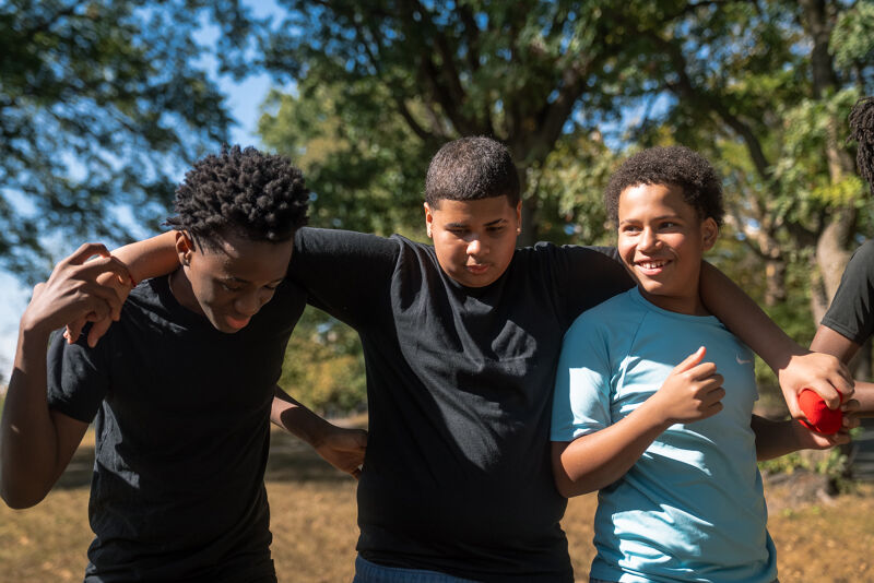 The image shows a group of young boys standing close together outdoors. They are holding each other, possibly as a sign of friendship or teamwork. The boys are of different ethnicities and are wearing casual clothes. The background is blurred, suggesting a park or natural setting.